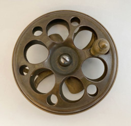 A brass side-mount reel. Large holes are cut through the sides to allow air circulation to help dry the silk lines used at the time.