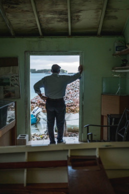 Ansil Saunders stands in his workshop doorway with his back to the camera, staring out onto the water.
