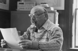 Joe Pisarro, seated, smokes a pipe as he examines a piece of paper.