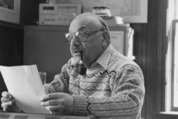 Joe Pisarro, seated, smokes a pipe as he examines a piece of paper.