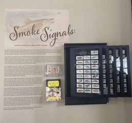 A photo of the Smoke Signals exhibition, featuring the cigarette card panels, a flattened cigarette box, and the book 