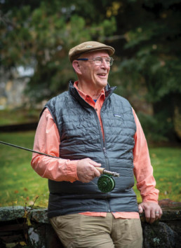 Paul Bruun, holding a fishing rod, leans against a stone wall.