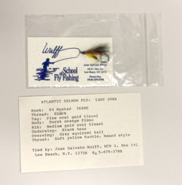 A fishing fly and business card for the Wulff School of Fly Fishing, featuring Joan Wulff's contact information, rest inside a small plastic bag. Underneath an index card lists the recipe for the fly, the Lady Joan.