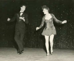 A man and a young Joan Wulff dance side-by-side onstage, their heads turned to look at each other.