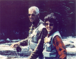 Lee and Joan Wulff turn to smile at the camera as they fly fish on a river.