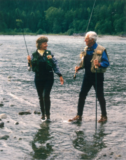 Joan and Lee Wulff hold fishing rods as they walk in the shallow waters of a stream.