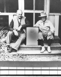 Two older men sit on steps in front of a building, a framing propped up between them.