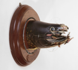 A carving of a brook trout from the gills to the nose. Various flies are hooked around the entirety of the mouth.