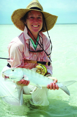 Nancy Zakon, crouches in shallow saltwater cradling a bonefish. She's wearing a long-sleeved pink shirt, white pants, and a straw hat.