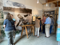 In the new Lee and Joan Wulff exhibition, a display case featuring an underwater movie camera and cans of film reels stands in front of a large image of Lee playing a fish. Visitors next to the case examine some of Lee's artwork that's hanging on the wall.