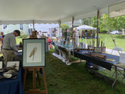 Tables arranged under a large tent showcase a wide array of offerings, including artwork, rods, and information about nonprofits.