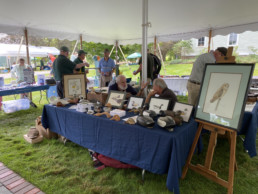 Two gentlemen in deep discussion sit behind a table displaying paintings and duck decoys in various stages of carving.