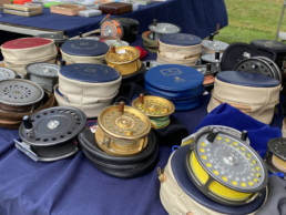 Dozens of fly reels, both with and without cases, are arranged on a table.
