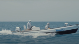 Two men speed across the ocean in a long flat boat named Daniella. Both are wearing light gray clothes, with hats and gaiters to protect them from the sun.,