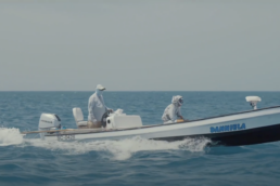 Two men speed across the ocean in a long flat boat named Daniella. Both are wearing light gray clothes, with hats and gaiters to protect them from the sun.,