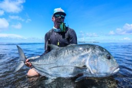 An angler, kneeling in the water, cradles a giant trevally. You can tell he is smiling widely even though part of his face is obscured by the fly reel and rod resting on his shoulder.