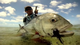 An angler displays a giant trevally; a large black streamer fly is hooked in its jaw.