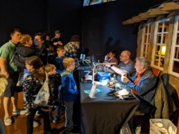 A group of kids and adults watch a woman and two men tie flies.