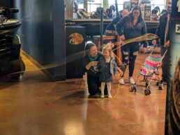 A woman kneels on the floor as she helps a young girl get a feel of fly casting. The girl's parents look on, smiling.