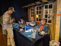 A woman and two men sit at a table tying flies while a man holding a travel coffee mug watches them work.