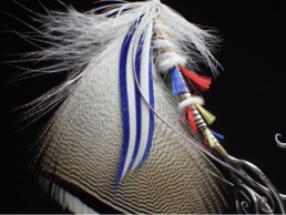 A closeup of an ornate, artistic fly. It has a wood duck feather wing, white hackle, a cheek of white and blue married feathers, and tufts of red, blue, and gold along its a white silk body.