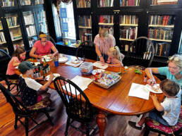 Four children and three adults gather in a library around a wood table. Most are seated and painting on paper plates.