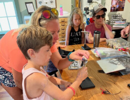 A woman and young boy are positioned in front of a fly-tying vise. He's holding on to the thread bobbin in his right hand, while she appears to be about to grasp the hook in the vice with her left thumb and forefinger.