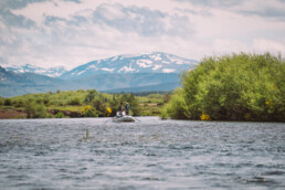 In the mid ground, three anglers on a float boat fish a river. While there are yellow flowers along the river bangs, a snow-covered mountain range is visible in the distance.