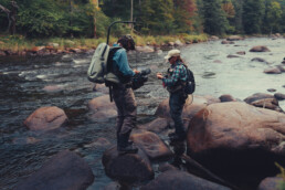 A cameraman and Rachel Finn stand on boulders in the middle of a river. He is focusing the camera on something in her hands.