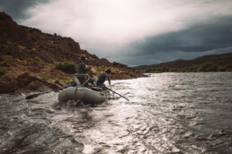 Three anglers in a float boat fish a river on an overcast day. One has a fish on, one uses oars to hold the boat steady, and one dips a long-handled net in the water.