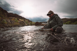 Rachel Finn crouches in a river, cradling a steelhead just at the surface of the water. It's a cloudy day, and she has a big smile on her face.