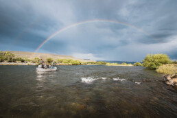 Three anglers on a river in a white motorized float boat. A double rainbow arcs over them.