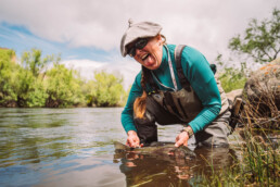 Rachel Finn crouches in the water, cradling a trout just at the surface. She's grinning with her tongue sticking out.