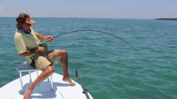Andy Mill, seated on a bench affixed to a boat, leans back as he attempts to reel in an unseen saltwater fish. Given the deep bend in his rod, it's clearly a sizable catch.