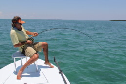 Andy Mill, seated on a bench affixed to a boat, leans back as he attempts to reel in an unseen saltwater fish. Given the deep bend in his rod, it's clearly a sizable catch.