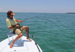 Andy Mill, seated on a bench affixed to a boat, leans back as he attempts to reel in an unseen saltwater fish. Given the deep bend in his rod, it's clearly a sizable catch.