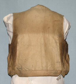 The back of a battered, much worn khaki fishing vest is displayed on an armless torso form. The sides in particular are stained with dirt and grime.