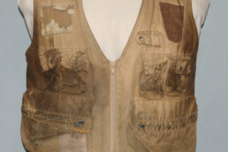 A battered, much worn khaki fishing vest is displayed on an armless torso form. The pockets and sides are stained with dirt and grime, and a patch, frayed edges, and a hole are visible in various spots.
