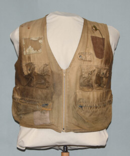 A battered, much worn khaki fishing vest is displayed on an armless torso form. The pockets and sides are stained with dirt and grime, and a patch, frayed edges, and a hole are visible in various spots.