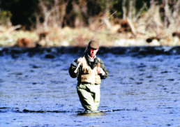 Prince Charles wades in the River Dee. His rod is propped on his right shoulder, and he's wearing fishing gear and a tweed driving cap. He's looking at the water with a slightly perplexed expression.