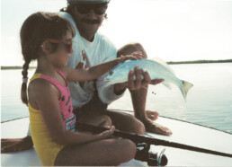 Jose Wejebe sits on a boat next to his daughter Krissy. He's holding a small fish in one hand, holding it towards her so she can tentatively touch its back. They're both smiling.