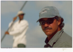 In the foreground, Jose Wejebe is turned to his left so he can look directly at the camera. He's wearing sunglasses and a blue-gray baseball hat and shirt. In the background is a blurry image of a guide holding a boating pole.