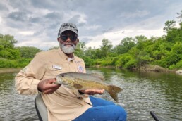 An older Black man with a gray beard sits on a boat cradling a bass to display for the camera.
