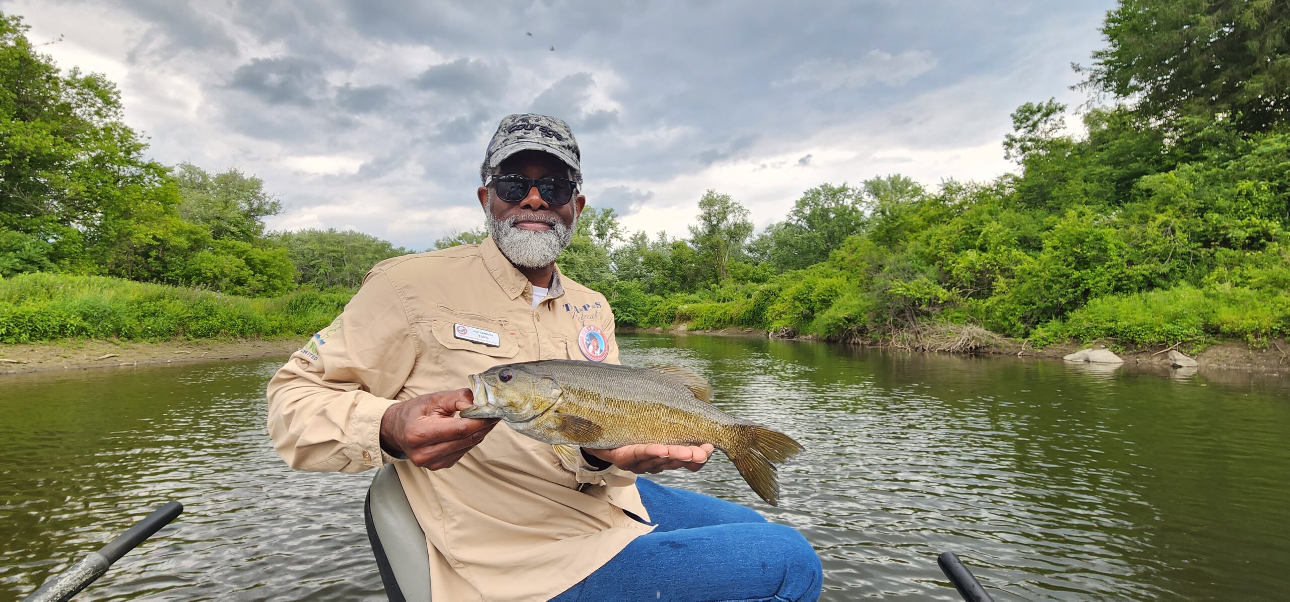 An older Black man with a gray beard sits on a boat cradling a bass to display for the camera.