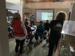 Steve Ramirez stands next to a projection screen in the darkened gallery. Several people sit in folder chairs as they listen to his talk.