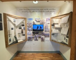 The main displays of the Jose Wejebe exhibition. A case on the left contains rods, reels, photos and a book, while the case on the right holds logbooks, calendars, and a storage box with flies. In the middle is a tv screen showing Jose Wejebe highlights; the screen is surrounded by photographs.