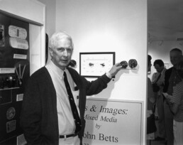 In this black and white photo, John Betts, an older white man wearing a cardigan, button-down shirt, and tie, poses with his left hand touching one of his reels.