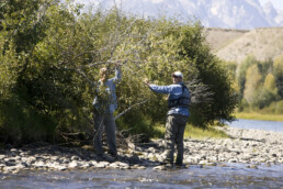 Two anglers, a man and woman, work to get their line untangled from brush at a river's edge.