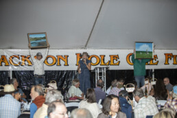 A blonde white woman in a denim dress, wearing a cowboy hat, stands on a stage and speaks into a mic. Two men flank her, each holding a painting up above their heads.