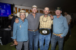 Four white men in casual clothes stand arm in arm. The second from the right is wearing an oversized belt buckle that spans the length of his waist; it features the Jackson Hole One Fly logo.